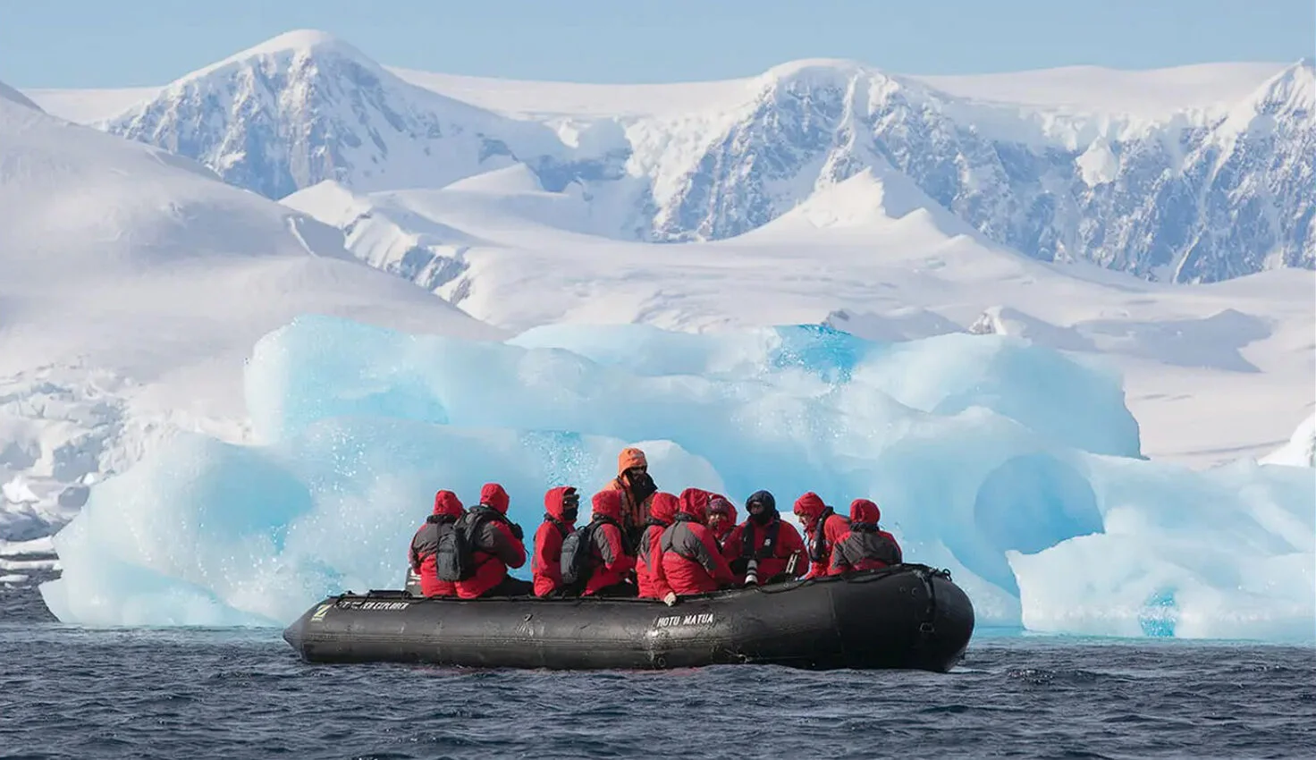 Pareja con tazas en las manos, apoyada en la barandilla de la cubierta, mirando un glaciar y montañas con una zódiac a lo lejos