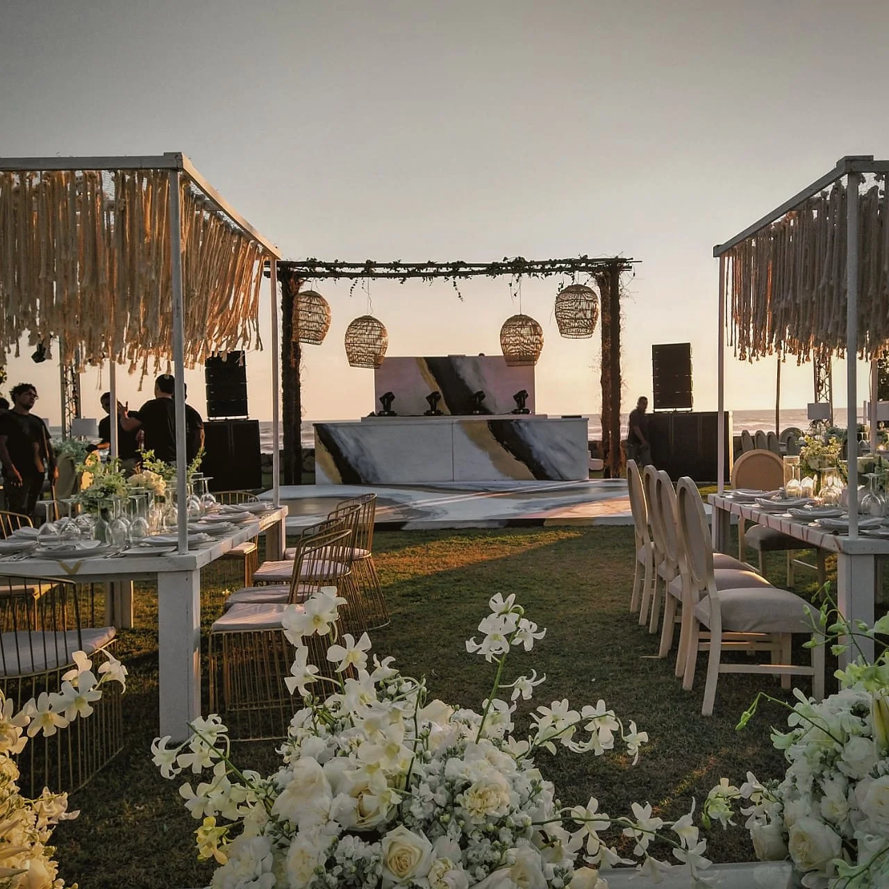 Montaje de boda frente al mar al atardecer, con mesas, cabina de DJ y audio coordinados con la planner