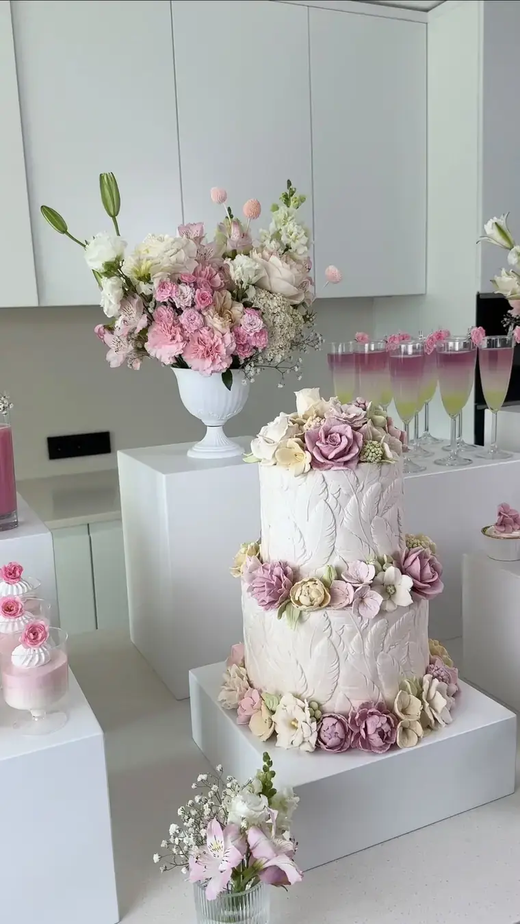 Two-tier white cake decorated with dusty pink and cream zefir flowers, beside fresh bouquets and champagne